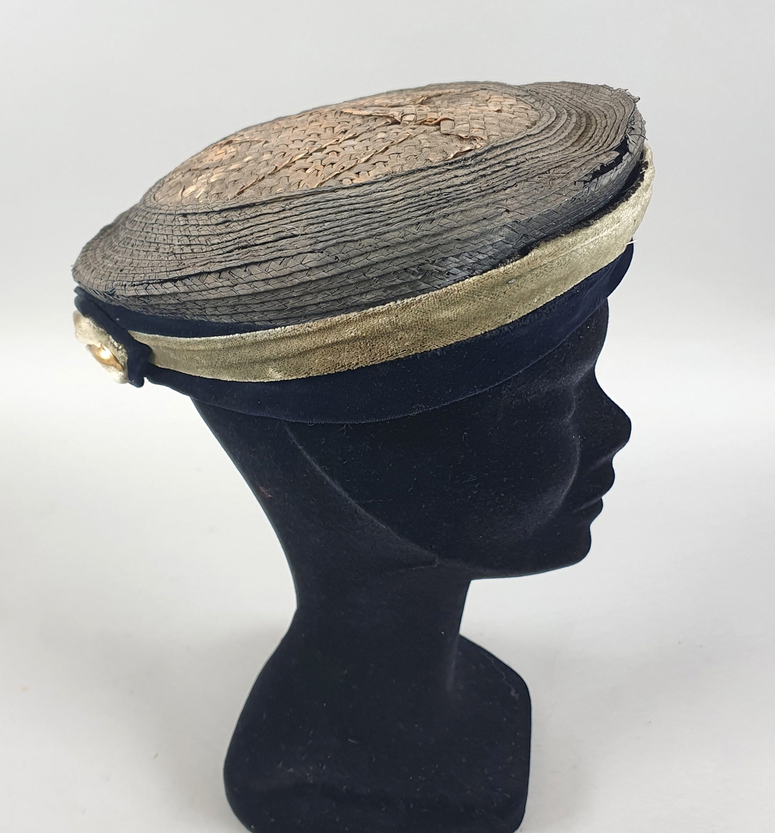 Rund og flat stråhatt med kant av grønn og blå fløyel og gul fløyelssløyfe midt bak. Kant på innsiden av skinn, som er løsnet fra hatten.