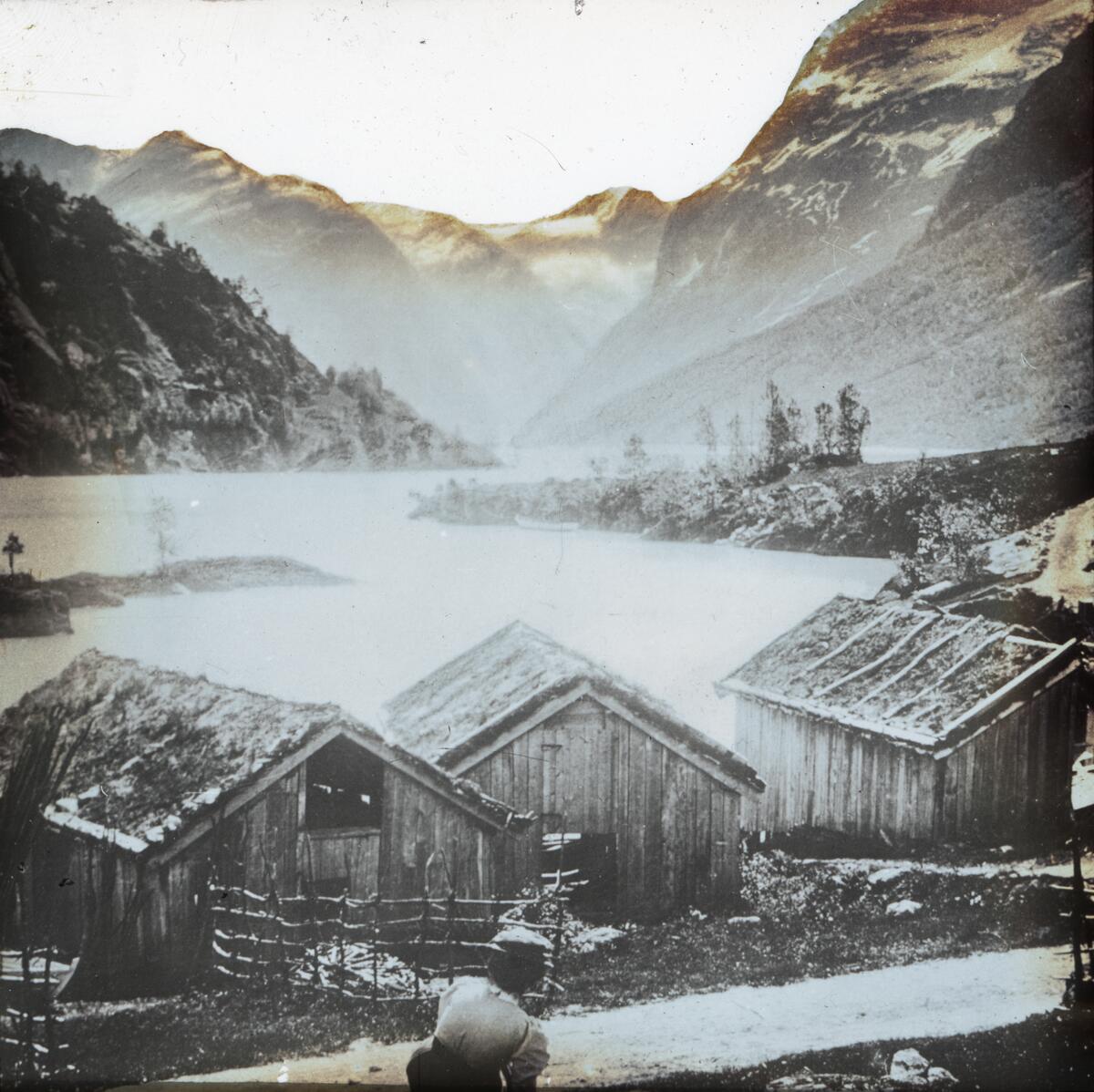 Dias. Tre naust eller uthus ligger ved Strynsvatn eller Loenvatn omgitt av fjell.