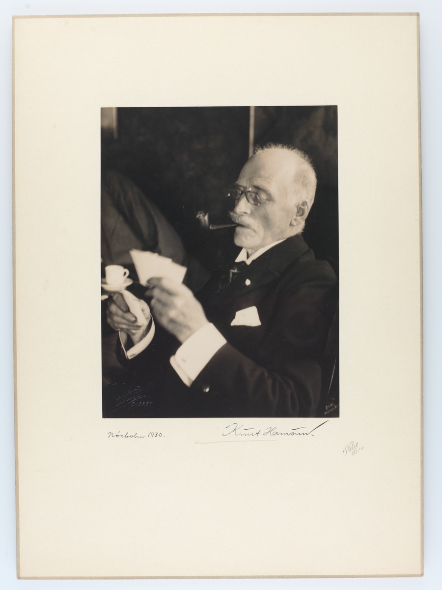 Portrett av Knut Hamsun med pipe i munnen og spillkort i hendene. Hamsun har signert bildet.