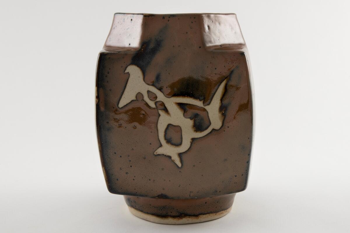 Vase med fire vegger og åttekantet munning.  Bygd opp i steingods glasert med persimonglasur (brun m. svarte spetter), og  penseldekor lagd med voksresist.  Dekor på alle fire sidene av vasen.