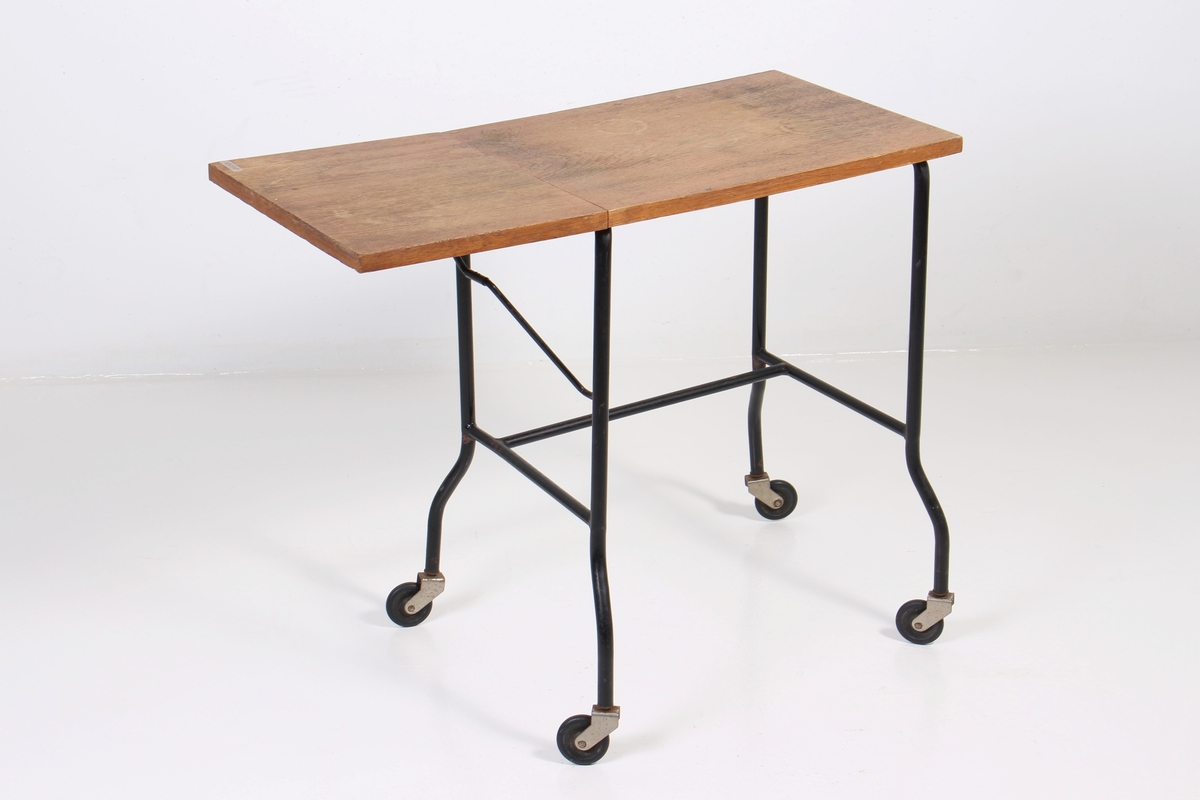 Lite avlastningsbord med finert bordplate, ramme av metallrør, og fire hjul. Bordplaten har en hengslet klaff som kan legges ned.