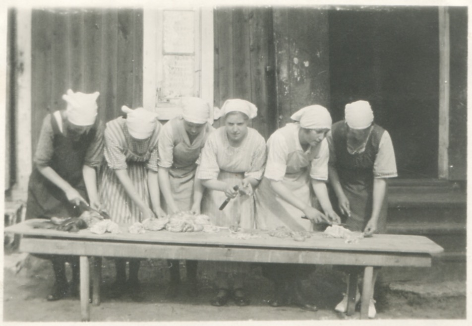 Sex kvinnor arbetar (med köttberedning?) utomhus vid ett långbord, "Nygård" Kållereds Stom 1:3 på Gamla Riksvägen, okänt årtal.