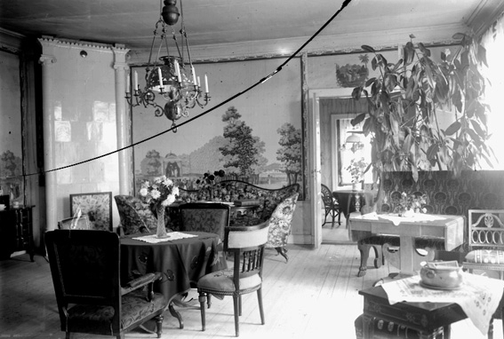 Salong med väggmålning och bord, stolar, prydnadssaker samt en lampa i taket.
Fotografens ant: Olssons, Höje.
