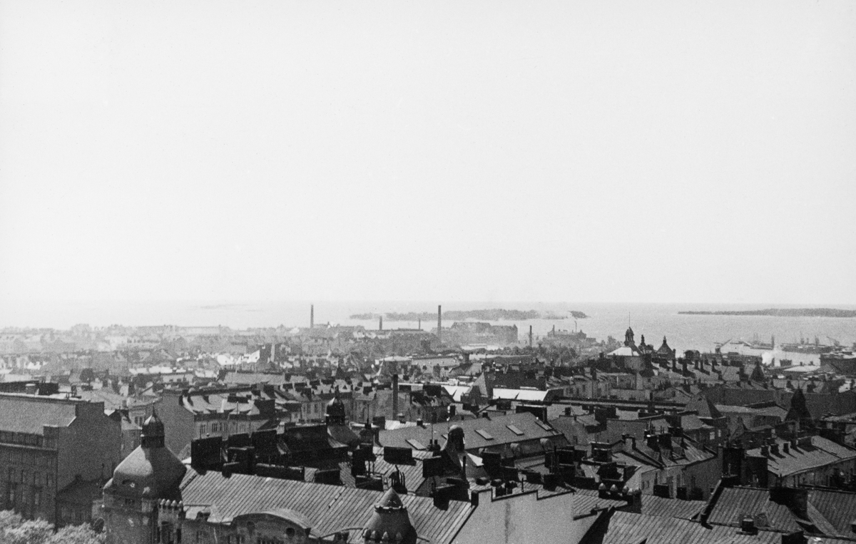 Utsikt over hustakene i den sentrale delen av den finske hovedstaden Helsinki eller Helsingfors, med havneområdet og et par av øyene i Finskebukta i bakgrunnen. Dette fotografiet ble tatt da norske fløtingsfunksjonærer var på studietur i Finland sommeren 1934.