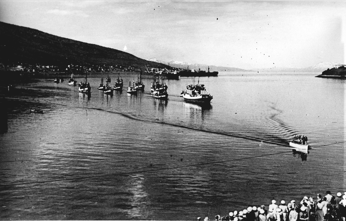 Båter på havna i Narvik. En sjalupp er på vei inn til folkemengde på kai i forgrunnen.