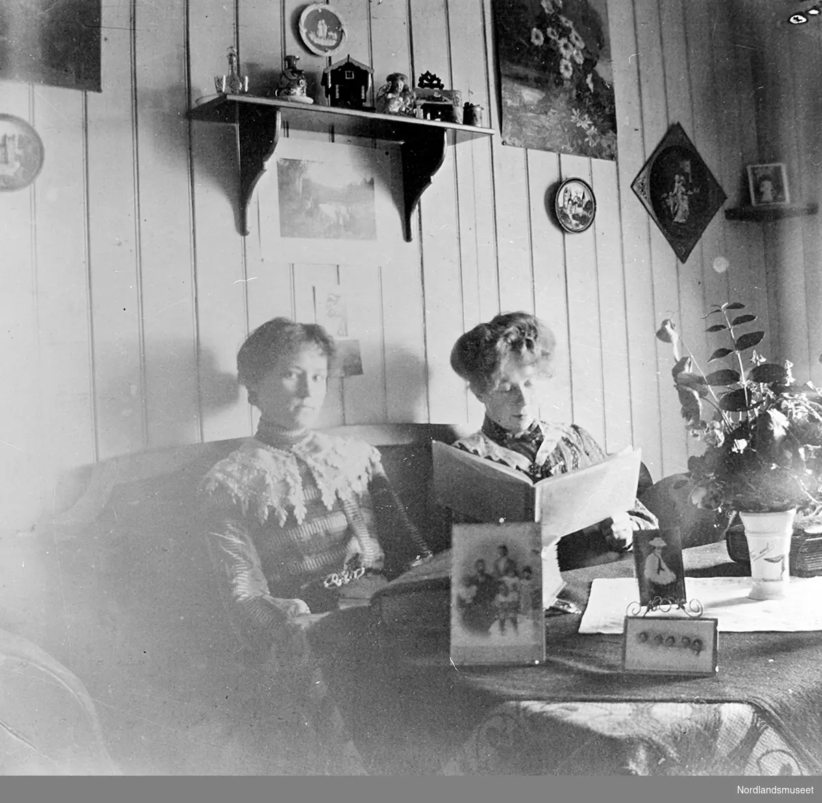 2 damer sitter i en sofa på hybelen i Lødingen, damen til høyre er Borghild Normann. På bordet foran en del fotografier og blomster. På veggen en hylle med små pynteting og en del bilder. Damen til venstre har strikket genser med stor lys krave, damen til høyre i mørk bluse med blondekrave. Hun leser i en bok.