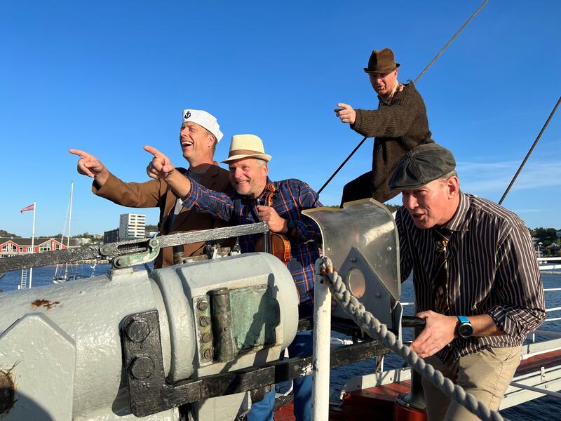 Fire menn står rundt en skipsharpun på en hvalfangstbåt. Alle ser mot venstre og to av mennene peker, de har skipsluer på seg. Alle smiler og ler på bildet.