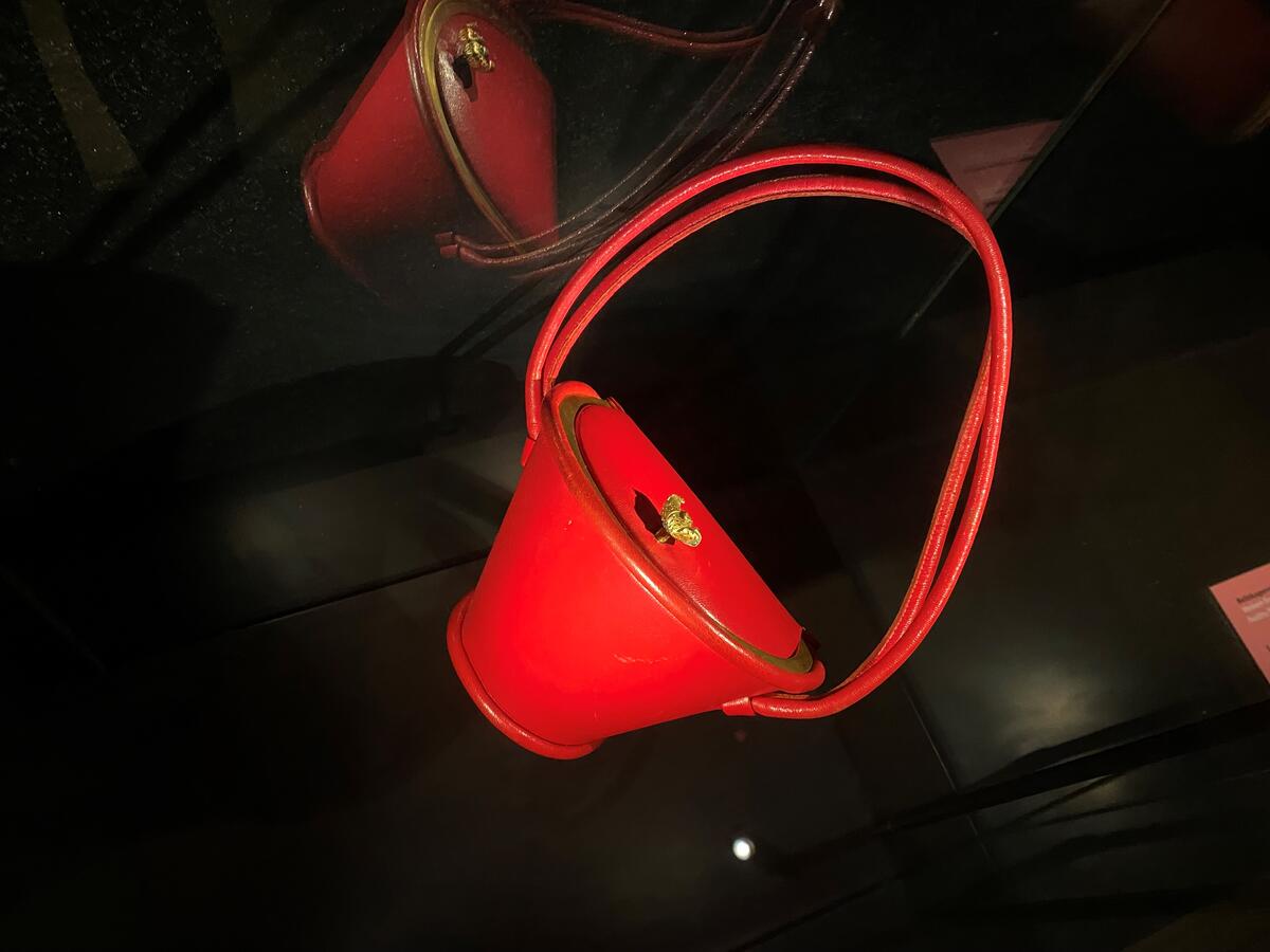Bildet viser en rød veske, den er traktformet med låsen i gull på lokket, har to remmer. Vesken er laget i selskinn.