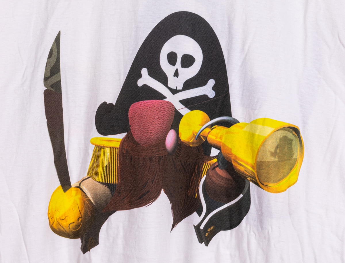Reklamtröja, vit t-shirt med tryckt bild av en stiliserad sjörövare på fram- och baksida. På ärmarna syns logotyper för olika tablettprodukter från Ahlgrens sortiment.
