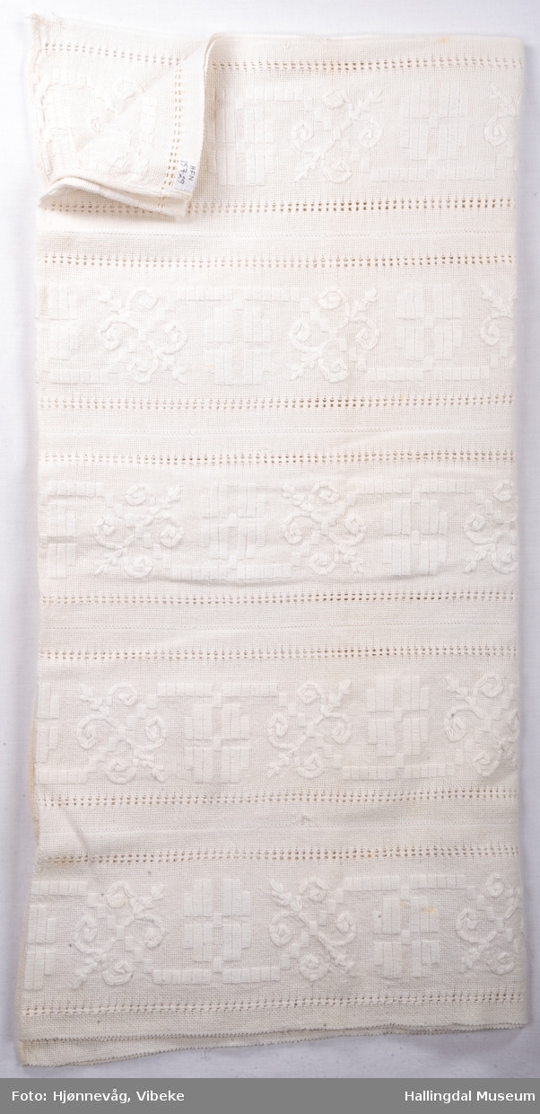 Bomullsduk vevd i toskaft, sydd sammen av 5 "remser" bomullstøy som hve er ca. 18 cm brede. Brodert med hvitt perlegarn.