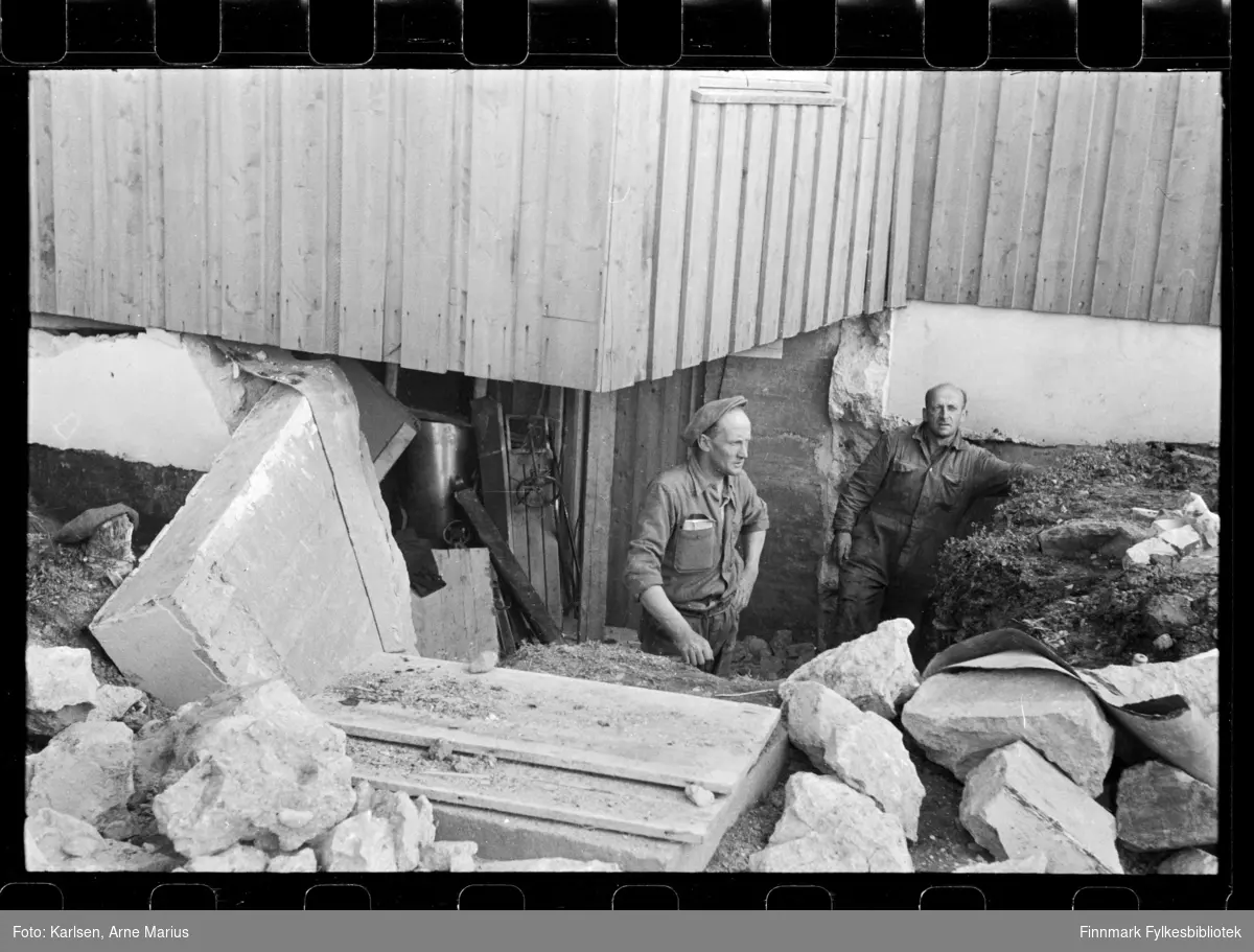 Skadet grunnmur på hus i Kirkenes 

To menn arbeider med reparasjoner

Foto antagelig tatt på slutten av 1940-tallet, tidlig 1950-tallet 