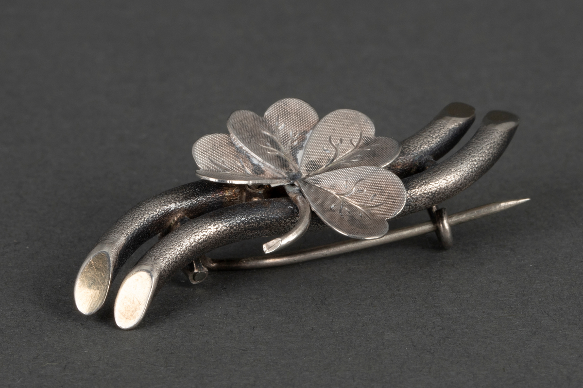 Nål/ brosje i sølv. Et firkløverblad er montert på to grenlignende rør. Gravyr på bladene. På smykkets underside er det festet en nål.