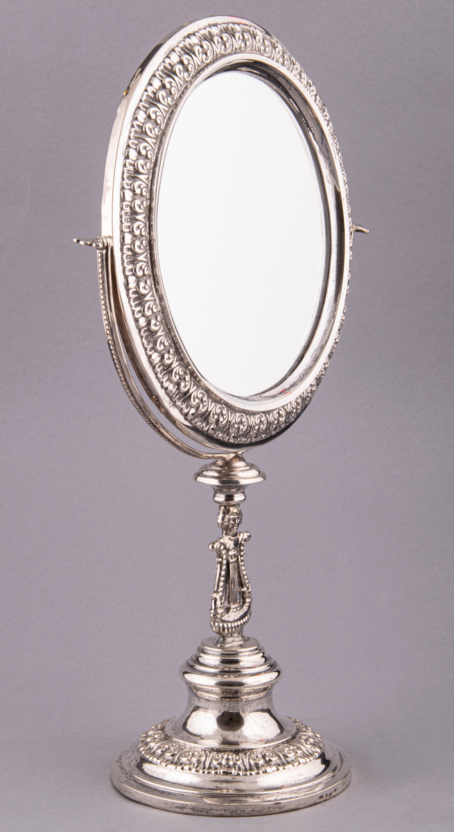 Bordsspegel i silver och glas. Tillverkad av silversmed Arvid Öhrn i Gävle, 1841. Rund på fot. Infattning m.m. av silver. Spegeln är vridbar runt i det halvcirkelformiga fästet, baksidan trä, teak? Rikligt med bladornament i kraftig relief.
I foten lyra med människohuvud med pagefrisyr. 
Stämplad "AÖ, G, L4"