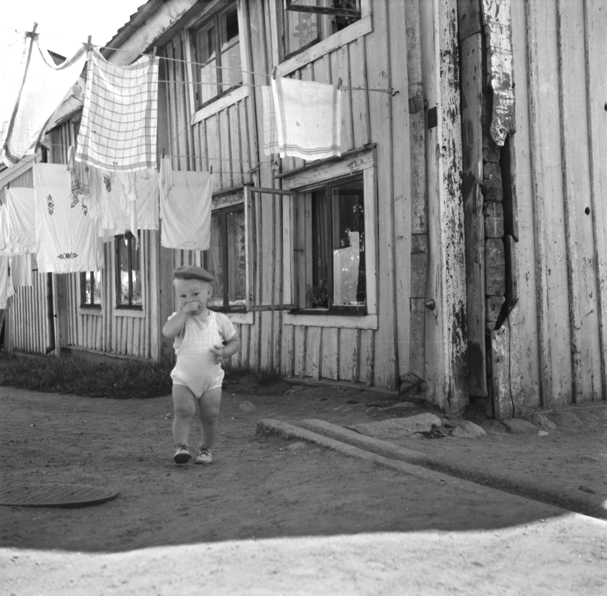 En gosse tultar fram bland tvätt på tork. Tvättstreck var förr en vanlig syn på stadens gårdar och utgjorde en uppskattad lekplats för lekande barn. Kvinnorna som svarat för tvätten delade oftast inte deras glädje. Pojken på bilden ska dock inte lastas, i alla fall inte i stunden. Foto från omkring 1950.