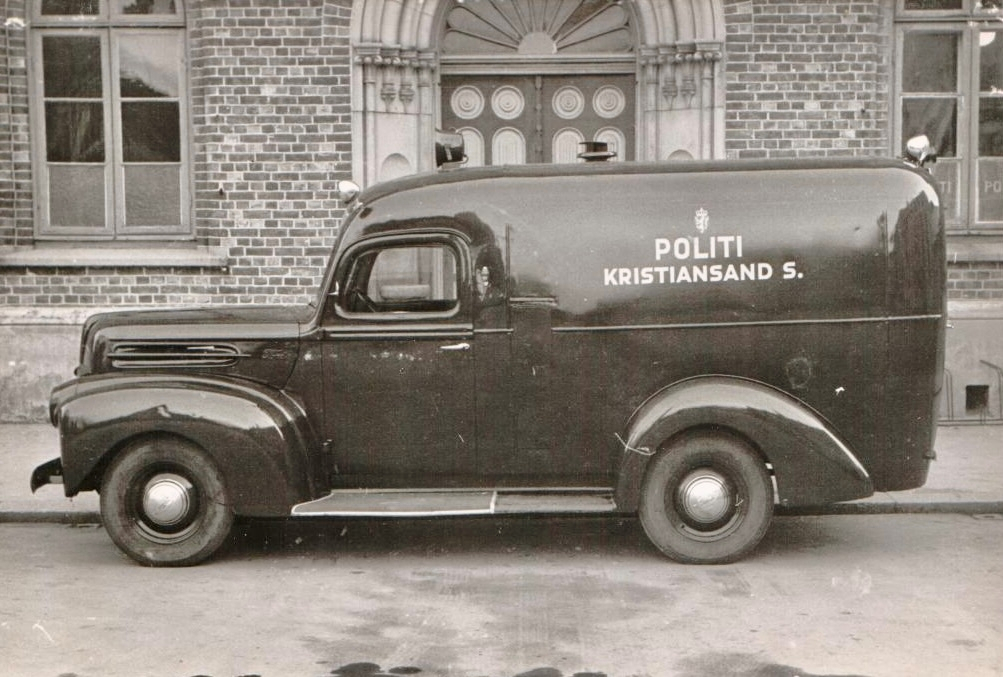 Cellebil, Ford 1946-modell, med registreringsnummer K-5 parkert foran en bygning. Bilen er merket "Politi Kristiansand S.", og er fotografert både forfra og fra siden.