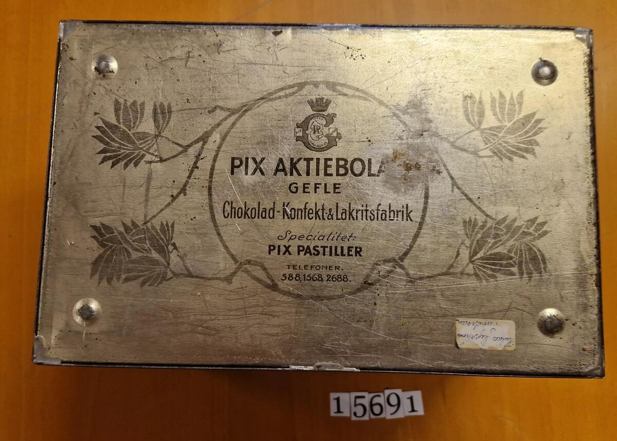 Plåtburk, formad som en kista. Pix Aktiebolag, Gefle. Choklad- Konfekt- & Lakritsfabrik. Specialitet: Pix Pastiller. Telefoner:588, 1568, 2688. Etikett i botten: Hulda Myrström, Sundsvall.