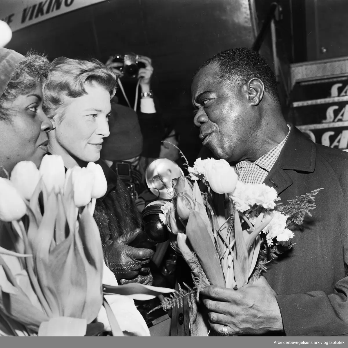 Louis Armstrong ankommer Oslo via Fornebu flyplass 2 februar 1959. Fra venstre: Lucille Wilson (hans kone), nrk-reporter Unni Børde og Louis Armstrong.
