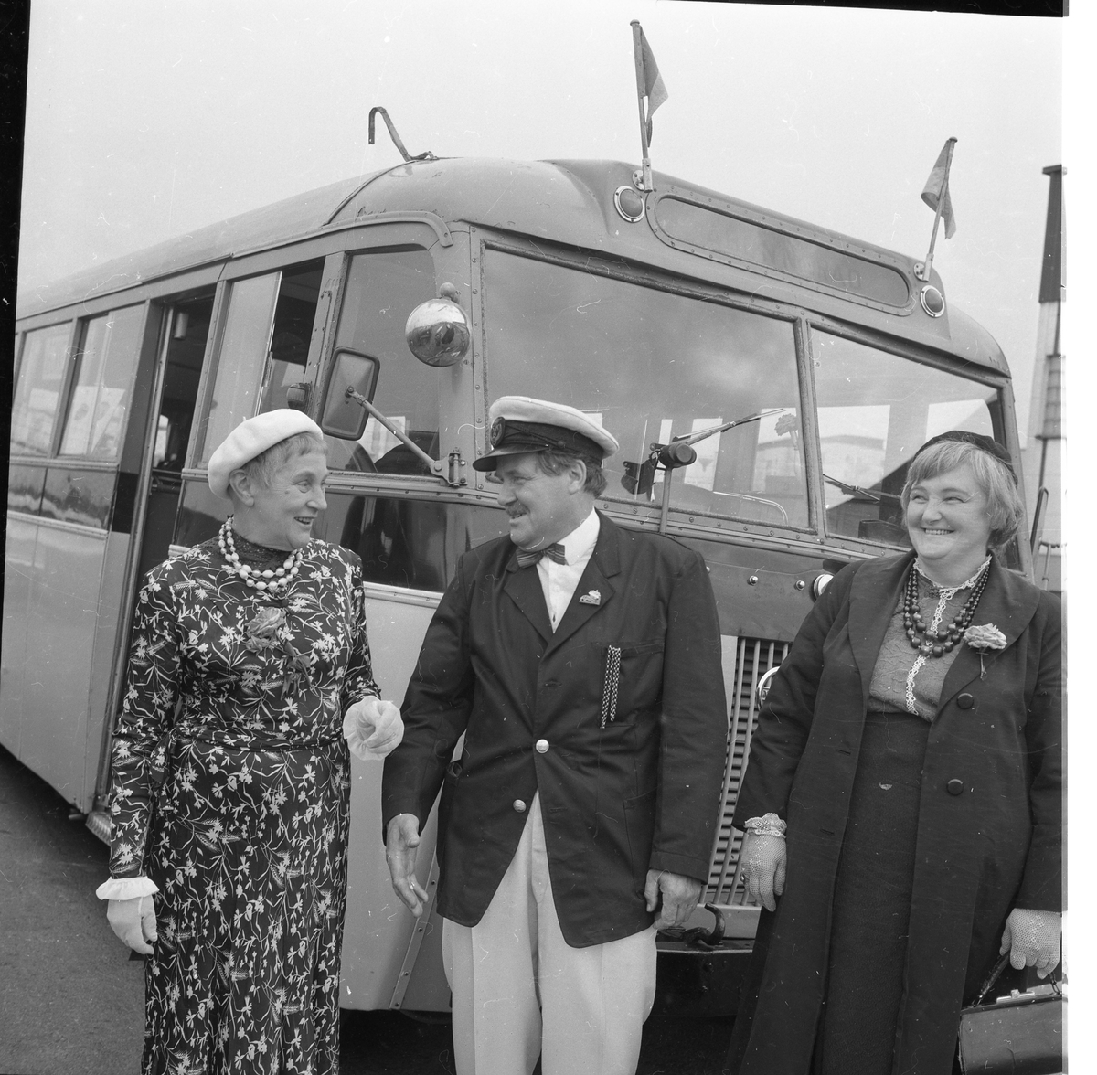 Två kvinnor flankerar en man som bär uniformskeps. De står framför en buss. De skrattar.