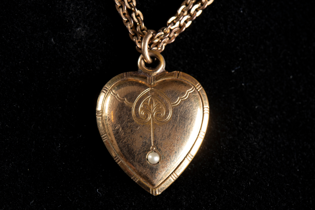 Halskedja i guldfärgad metall med hjärtformad berlock. Berlockens framsida graverad och ornerad samt försedd med en infattad pärla.