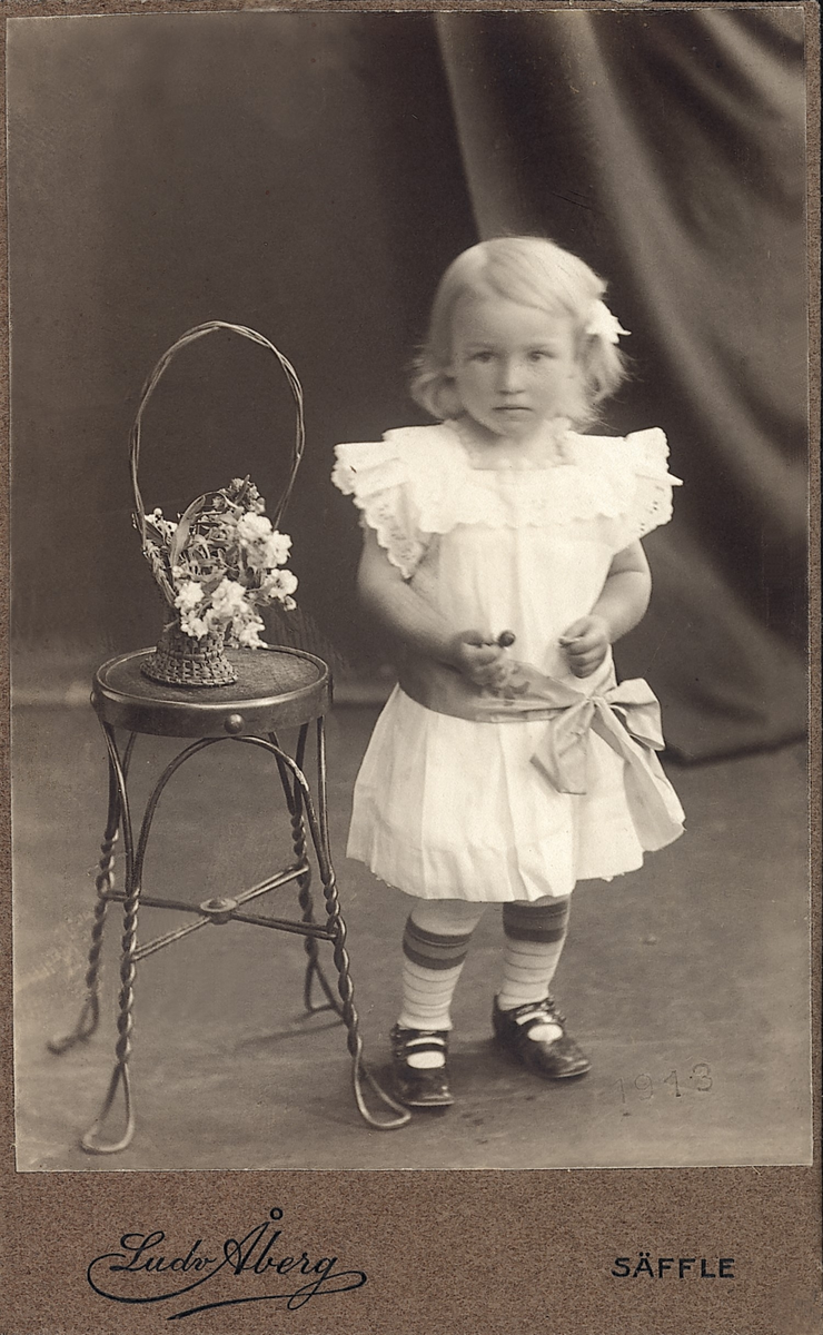 Ateljéfotografi av en liten flicka, stående bredvid en pall med en blomsterkorg på.