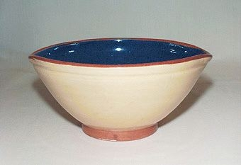 Gul skål av lergods försedd med två pipar, blå insida samt brun kant och fot av transparent glasyr.