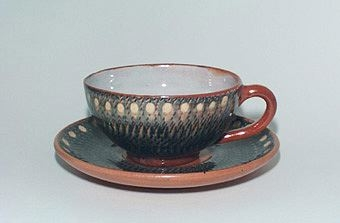 Kaffekopp med fat av lergods. Koppen är invändigt glaserad i grått och har ett rödbrunt öra. Koppens utsida är blågrön, gul och rödbrun med hämrat och hornmålat mönster. Likaså är fatets utsida, men undersidan är rödbrun med transparent glasyr.