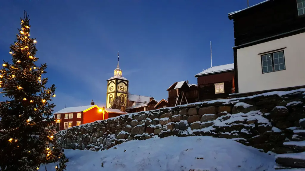 Bygninger, en steinmur og ei kirke. Foran er et juletre. Det er kveld/natt.