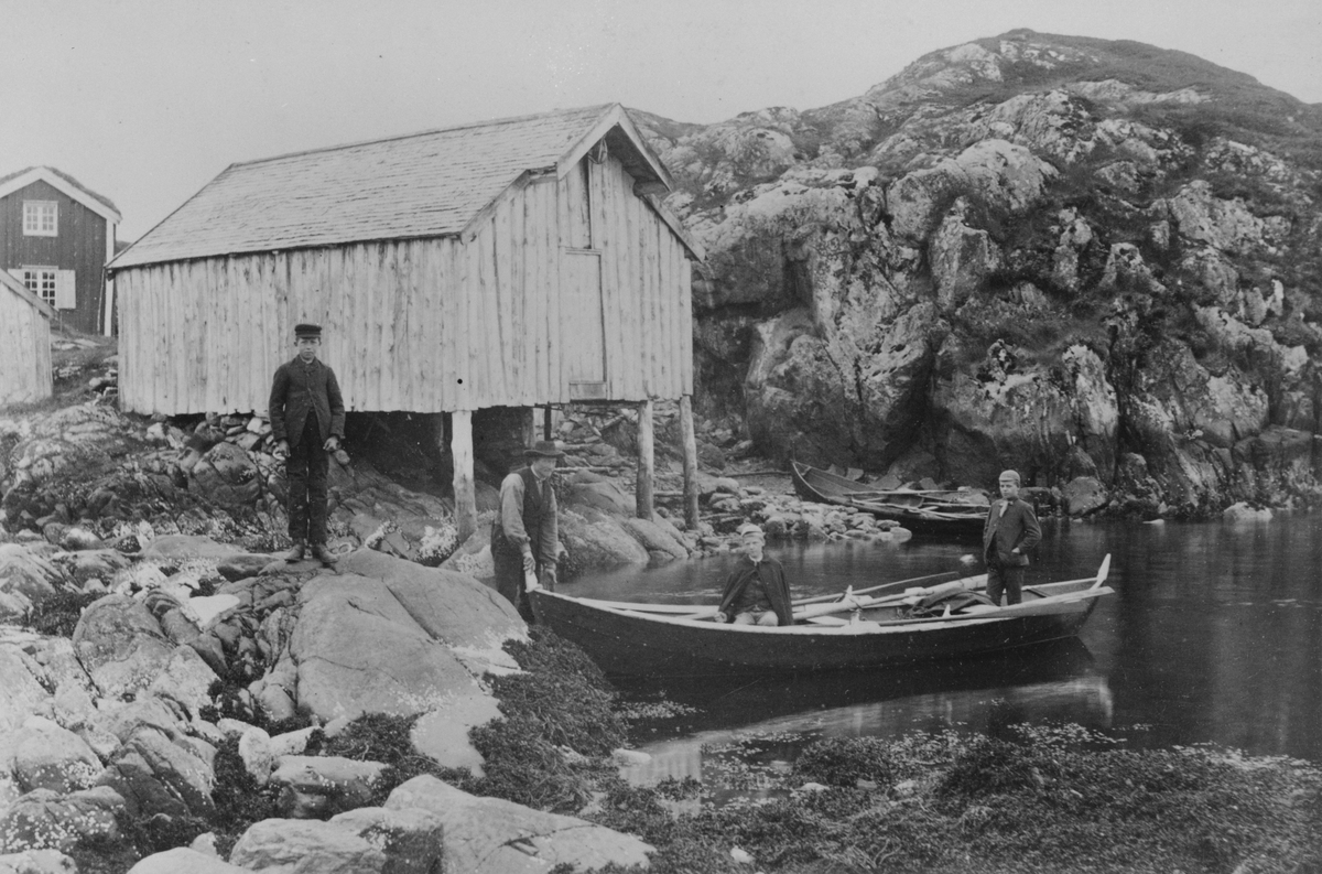 To gutter i en robåt på vannet ved et naust. En gutt til står på land, samt en mann som holder i båten.