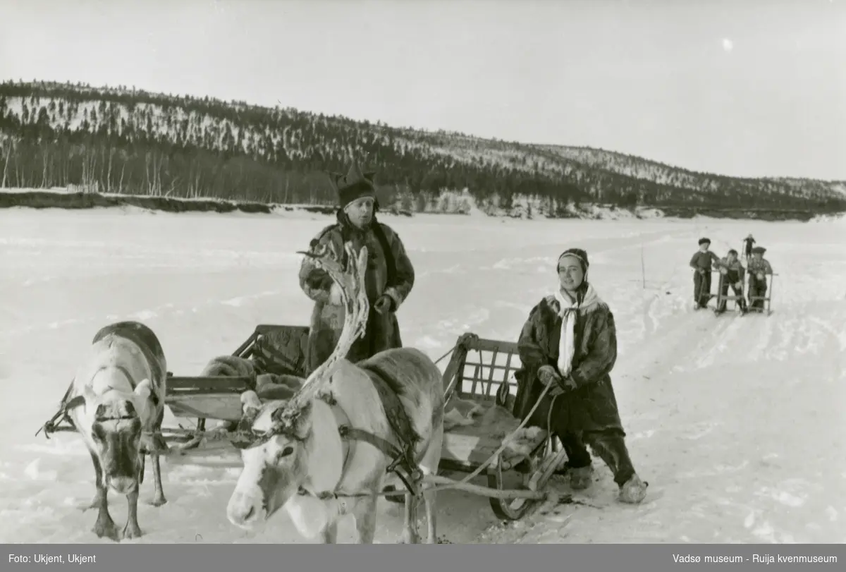 En flott vinterdag med rein, sleder og en kvinne og  mann i pesk, skaller og samiske luer. Bak kommer 4 barn på spark.