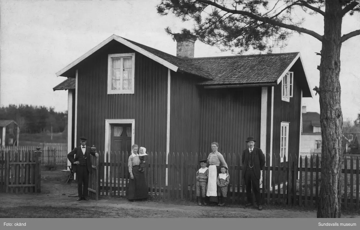Två kvinnor, två män och tre barn står invid staketet framför ett rödmålat bostadshus på landsbygden. Ur ett album från Matfors.