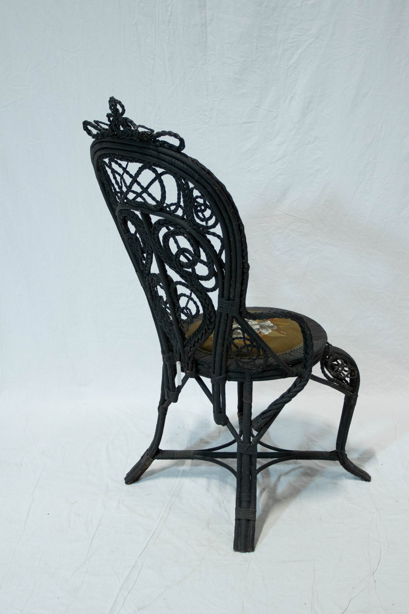 Svart kurvstol. Stolen har bøyde vidjeknipper som konstruktsjons ledd og et kunstferdig utført fletteverk av ornamental virkning i stolryggen. Setet er overstoppet med broderte perler og petit-point.