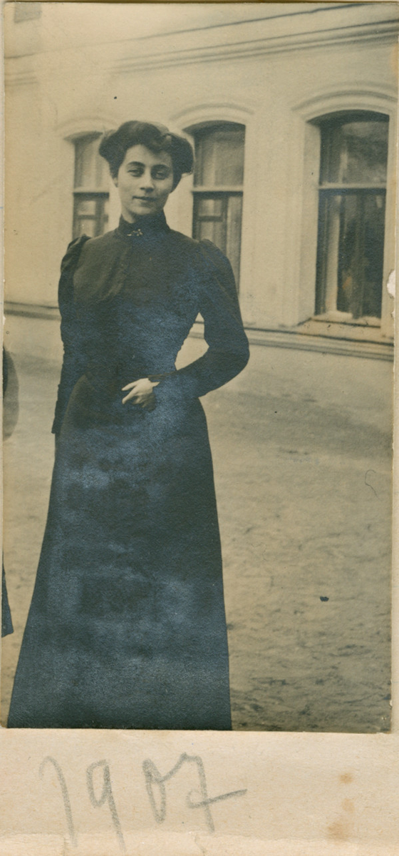 Florentine Rostin fotografert utendørs i 1907. Hun står på gaten foran en bygning, ikledd en mørk kjole.