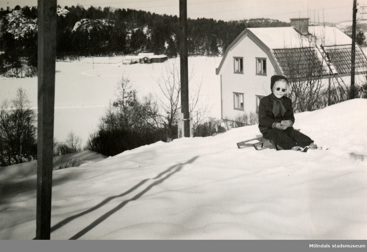 Eva Pettersson (född 1944, gift Kempe) sitter på en kälke framför bostaden på Gamlehagsvägen (ej i bild) år 1951. I bakgrunden ses Alvered.
