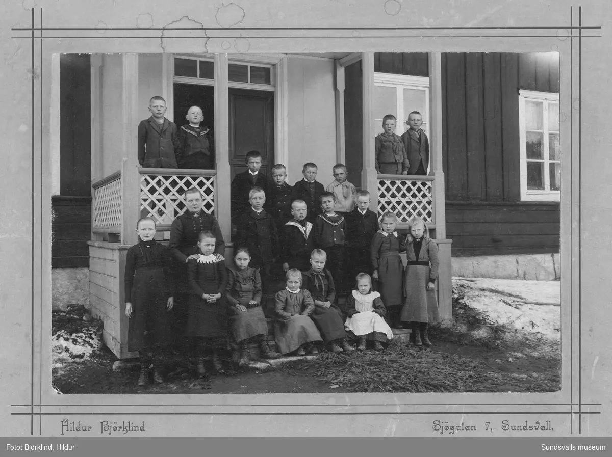 Skolklass och lärarinna på brokvisten till en träbyggnad. På baksidan av bilden står: Frida Ottilia Mikaelsson, Målsta. Ur Anders Nylanders samling.