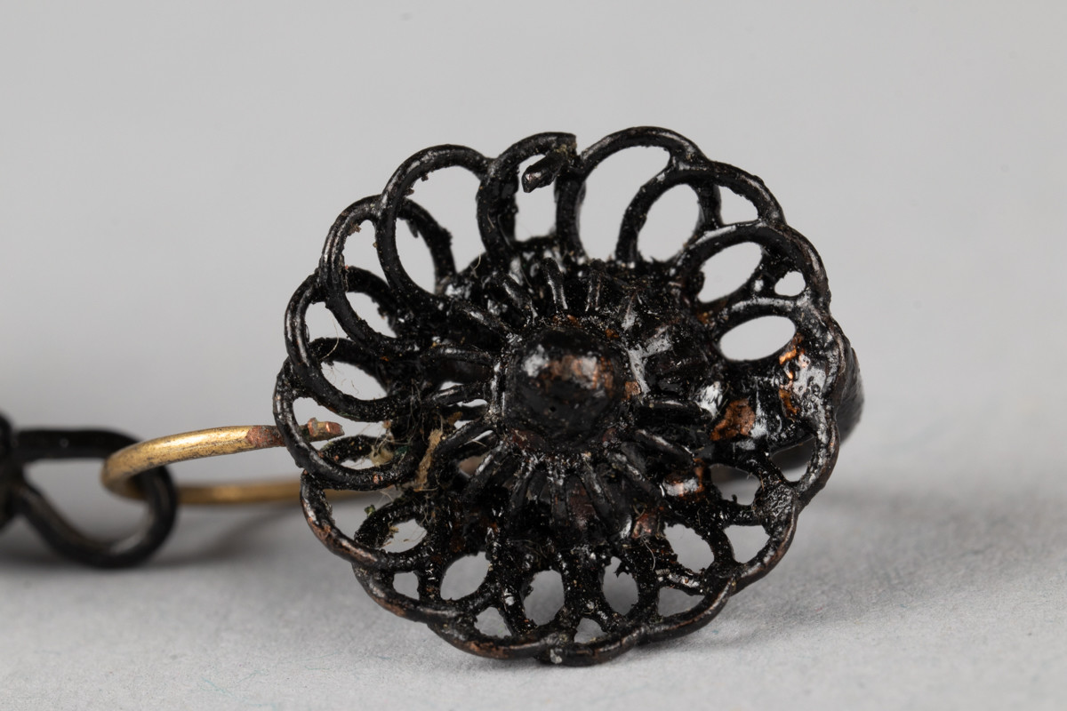Dråpeformede ørepynt med en rosett på hekten til øret og mørke metall tråder som danner dråpeformen under.
