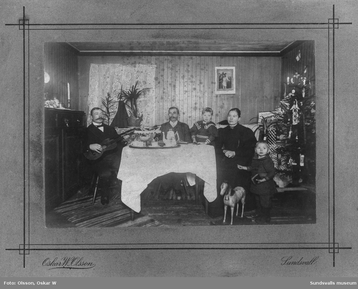 Två män, en kvinna och två barn vid ett bord med dukad kaffebricka. En av männen knäpper på en gitarr. En pyntad julgran står vid ena sidan. Ett av barnen har en stor leksakshäst i trä. På baksidan står: Familj i Torpshammar. Ur Anders Nylanders samling.