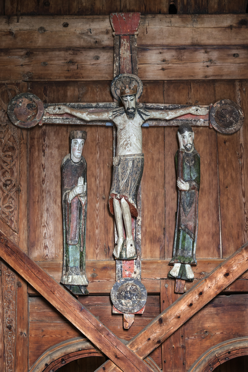 Kalvariegruppe skåret i tre, malt. Kristus på korset med krone, nagler gjennom hender og føtter. Maria tilvenstre, Johannes tilhøyre. Farger: lys grålig hud, grønne og rødlige farger.