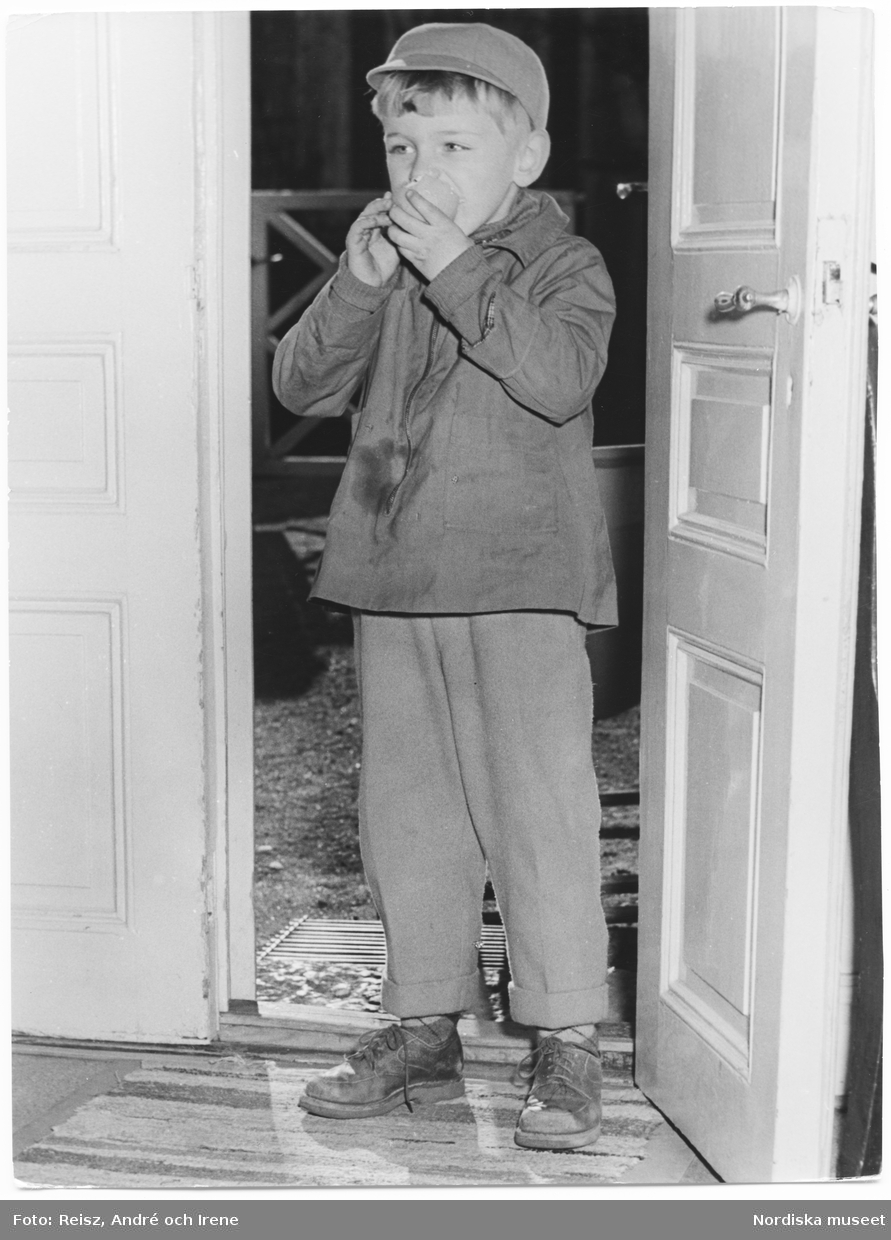 Helporträtt av en pojke som står i dörröppningen mot gården och äter ett äpple.