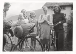 Tre kvinner og tre unger/ungdommer med to sykler foran huset