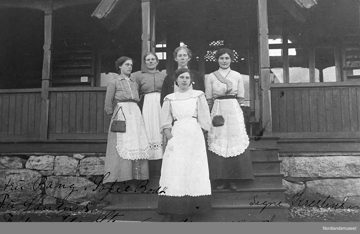 Betjeningen ved den gamle turisthytta, Bodø, Rønvikfjellet. 5 kvinner står på trappen til turisthytta. Fremst fru Bang. Ellers fra venstre: Sofie Bodø, fru Sandnes, frøken Ida Strand og Sofie Kristensen. Alle har hvite forklær på seg.