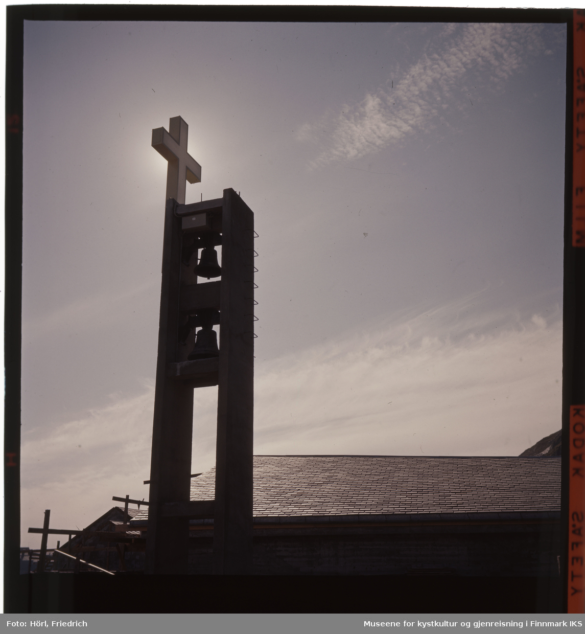 Kirketårnet til Den katolske kike St. Mikael i Hammerfest med kors og klokkene er fotografert i motlyset. Tydelig ser man kirketaket som er tekket med rektangulært takskifer. Til venstre for bygget står det fortsatt noe stilas.