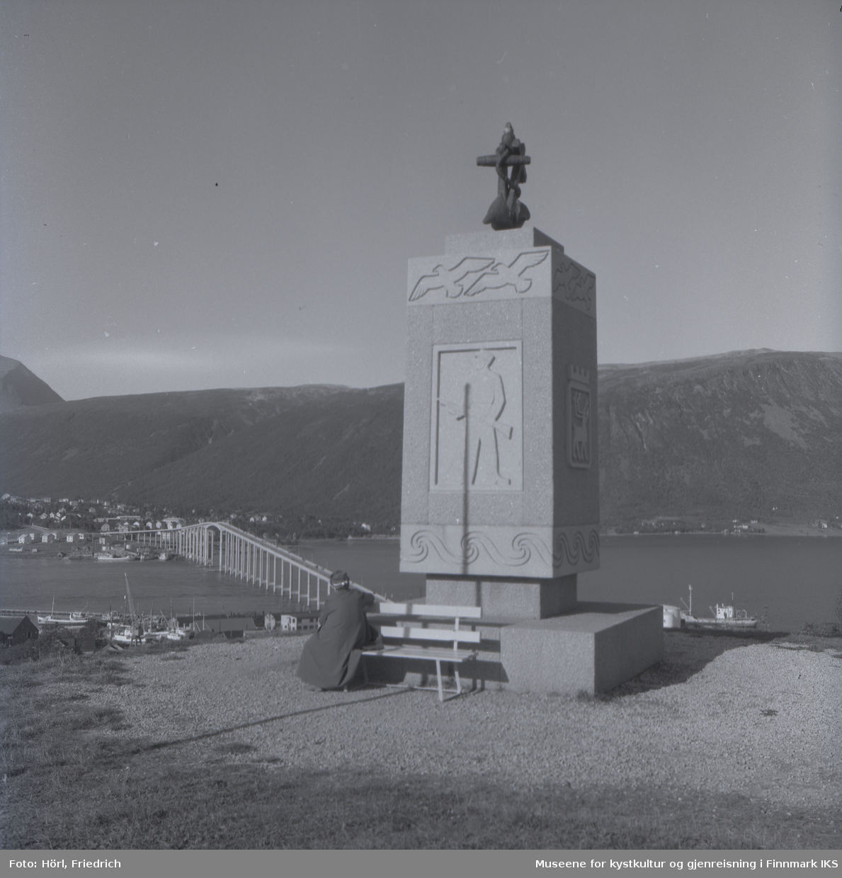 Monument ved "Tromsø Maritime skole". En kvinne sitter på en benk og ser ut over Tromsøysundet. I bakgrunnen ser man Tromsø-brua, fjell og bebyggelse.