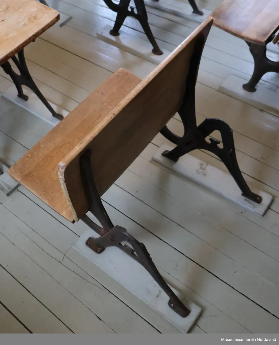 Skolepult med svungne bein av støpejern skrudd fast i to planker, samt sete med ryggplate av tre. Under bordplaten er et rom. Setet kan vippes opp og ned.
