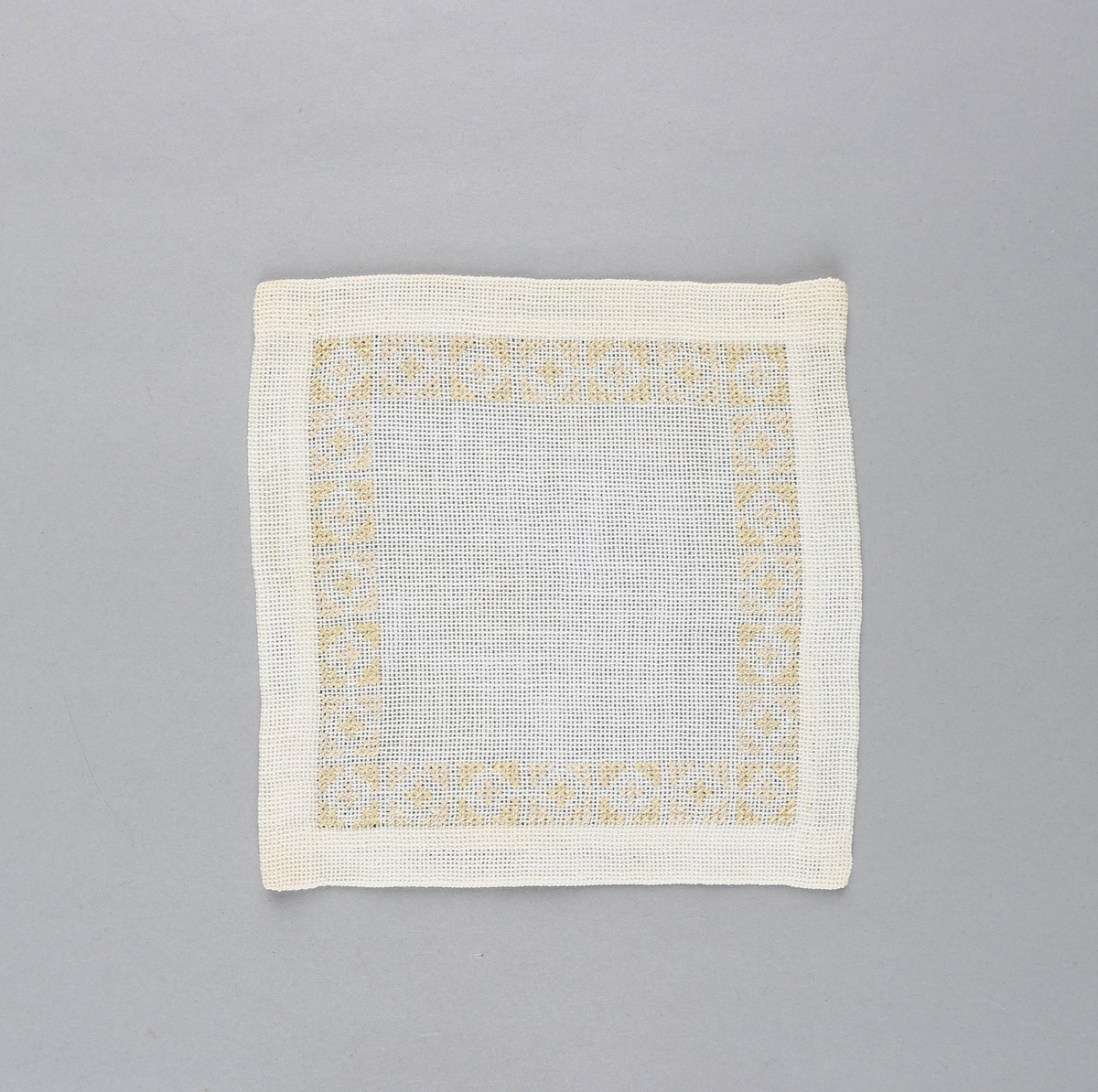 Tilnærmet kvadratisk dekkebrikke i hvitt tekstil med brodert bord langs brettekanten. Broderiet består av korssting av gul og rosa tråd i geometrisk mønster.
