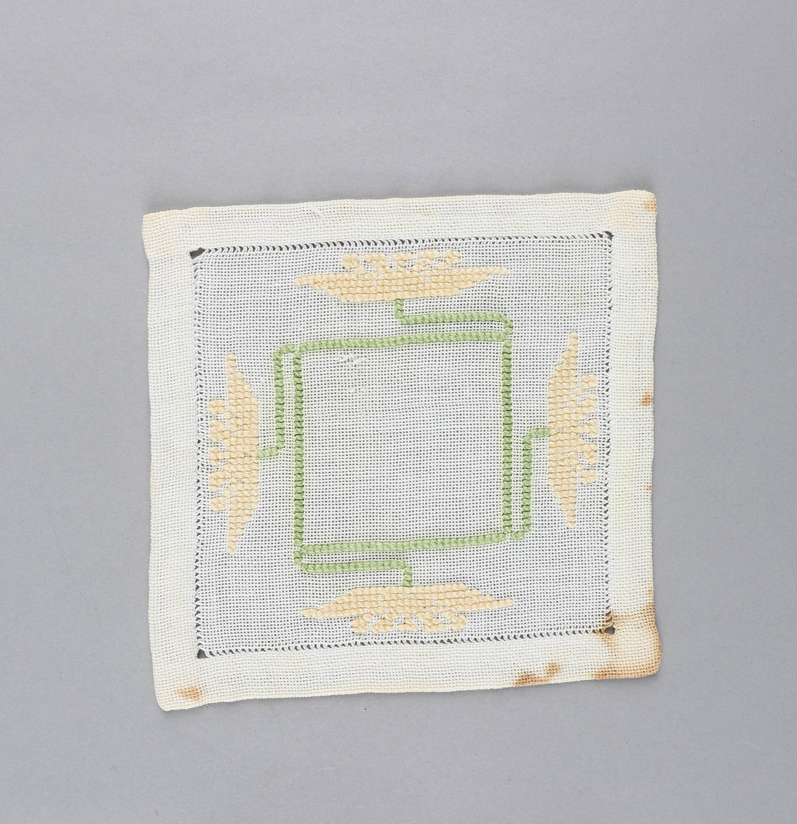 Tilnærmet kvadratisk dekkebrikke i hvitt tekstil med brodert, grønt kvadrat med fire stilker i sidene med gule blomster i endene.
