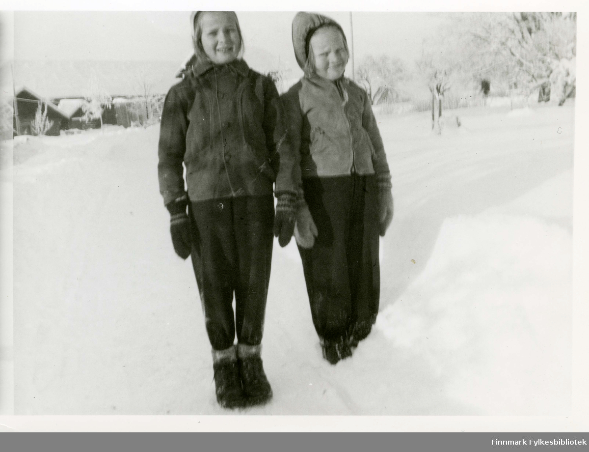 Eli Alexandersen med sin søster Berit (til venstre) i jakke som kom i Amerikapakke, "Marshallhjelppakke". Jakken var ny da den kom. Vindstoff utenpå og foret med rødrutet ullstoff. Glidelås på hetta, kunne brettes ned til krave når den ble åpnet. Pakken kom i 1945 da Eli var 7 år. På bildet har Berit på seg jakken, hun er 3 og et halvt år yngre enn Eli.  Bildet er tatt i Trøgstad der familien bodde under evakuering.