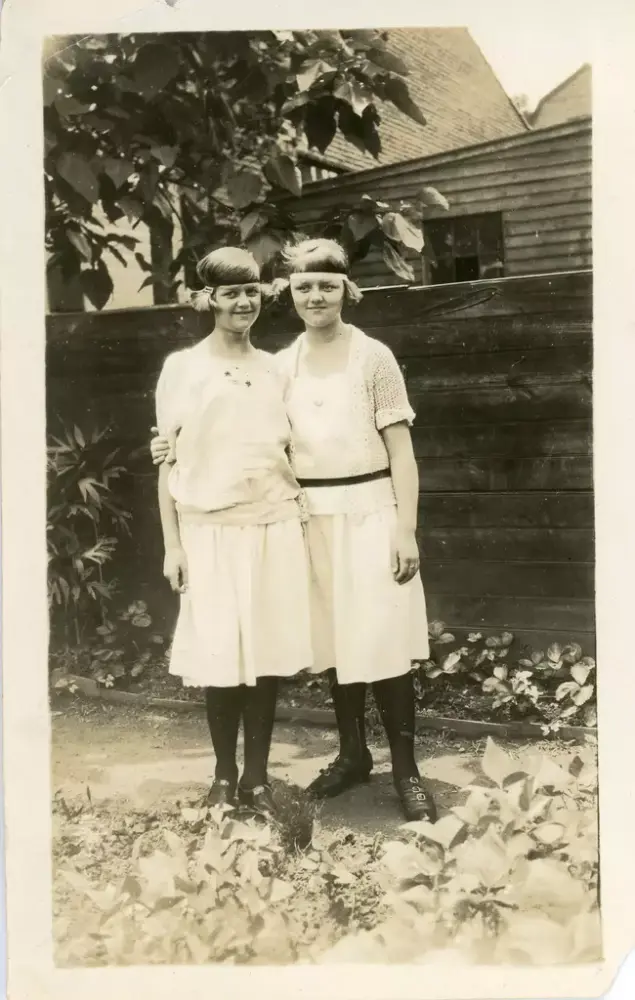 Gammelt foto av to unge jenter. De står ute. De har kort hår. Klesstilen og hårbåndet minner om flapperstilen på 20-tallet. Den ene jenta har hånden rundt den andre.