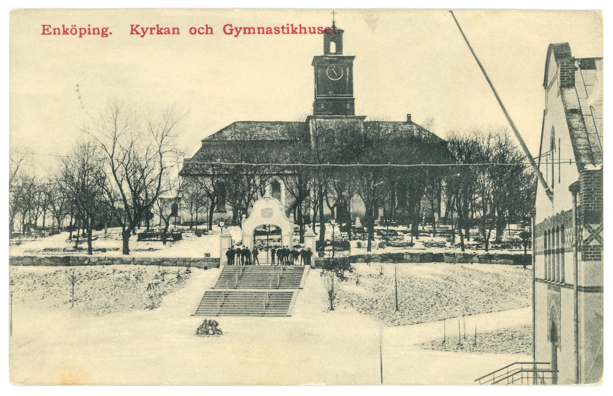 Vykort med motiv av skolpojkar i trappan framför Vårfrukyrkan och dess portal. Till höger i bild syns lite av gymnastikhuset, Enköping. Vykortet är poststämplat den 2/5- 1912.