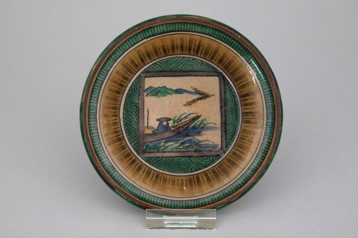 Skål med flatt speil. Utover rundede sider. Helt dekket av emaljemaling i gult, grønt, blått, brunfiolett og sort over grå glasur. Motivet viser fisker i båt, "fuku" i form av seil.