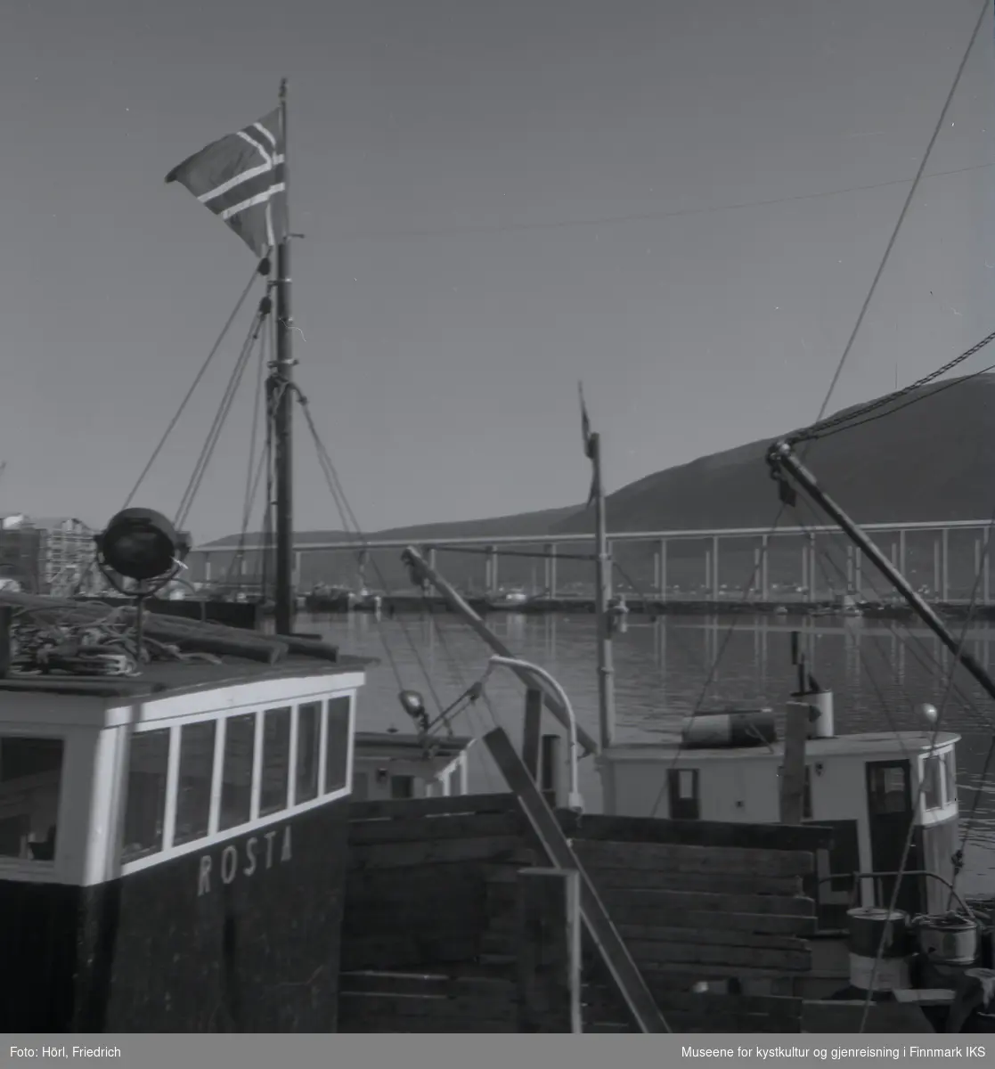 Skuta "Rosta" og flere fiskebåter bak den, til kais i Tromsø. Et av båtene har heist det norske flagget i masta. I bakgrunnen ser man Tromsøbrua.